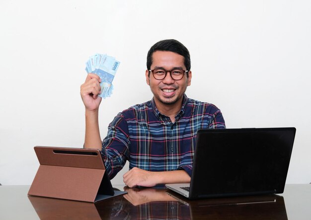 Asiatischer Mann lächelt, während er Geld vor seinem Laptop und seinem mobilen Tablet zeigt