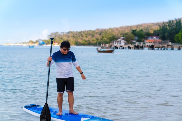 Asiatischer Mann mit Brille in Freizeitkleidung, der auf dem Paddleboard steht, Mann glücklich Paddle-Boarding auf blauem Meerwasser im Sommerurlaub in Thailand Island Beach