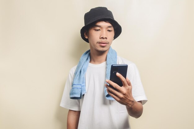 Asiatischer Mann mit hellweißem T-Shirt in schwarzem Hut mit blauem Handtuch und Telefon auf isoliertem Hintergrund