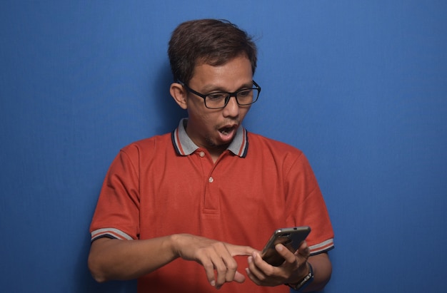 Asiatischer Mann mit orangefarbenem Casual-T-Shirt mit Smartphone mit Überraschungsgesicht und erstauntem Ausdruck