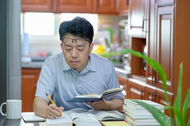 Asiatischer mann mittleren alters, der zu hause am schreibtisch sitzt, ein buch liest und studiert.