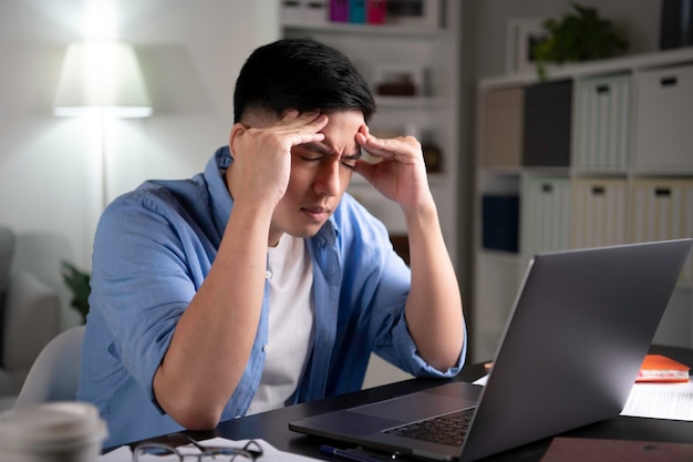 Asiatischer Mann müde von harter Arbeit mit geschlossenen Augen mit Laptop, um von zu Hause aus zu arbeiten