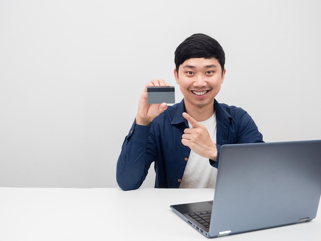 Asiatischer Mann sitzt mit Laptop am Tisch und zeigt mit dem Finger auf die Kreditkarte in der Hand