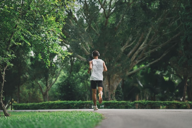 Asiatischer Mann Sportler fitter Körper mit weißen Kopfhörern und intelligenter Uhr beim Laufen oder Joggen im Garten haben mehr Baum und saubere Luft beim Trainieren im Park Gesundes Laufkonzept für Sport