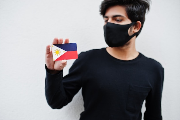 Asiatischer Mann tragen ganz schwarz mit Gesichtsmaske halten Philippinen Flagge in der Hand lokalisiert auf Weiß. Coronavirus-Länderkonzept.