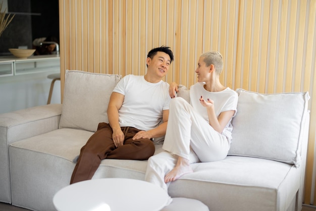 Asiatischer Mann und kaukasische Frau, die zu Hause auf dem Sofa sitzen und sprechen. Konzept der Beziehung und gemeinsame Zeit verbringen. Idee des häuslichen Lebensstils. Lächelndes gemischtrassiges Paar im Studio-Apartment