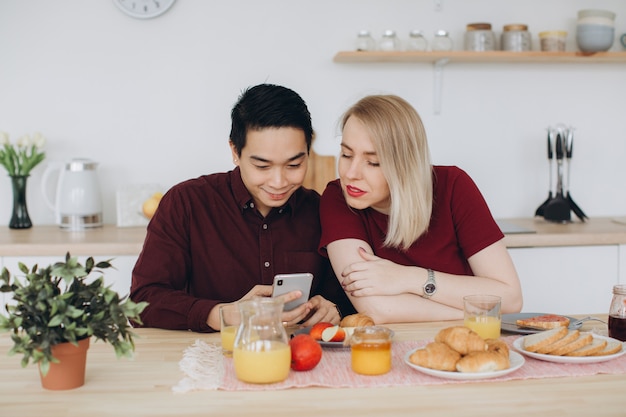 Asiatischer Mann und seine kaukasische blonde Frau frühstücken. Sie verbringen Zeit miteinander in der Küche und schauen sich Videos am Telefon an.