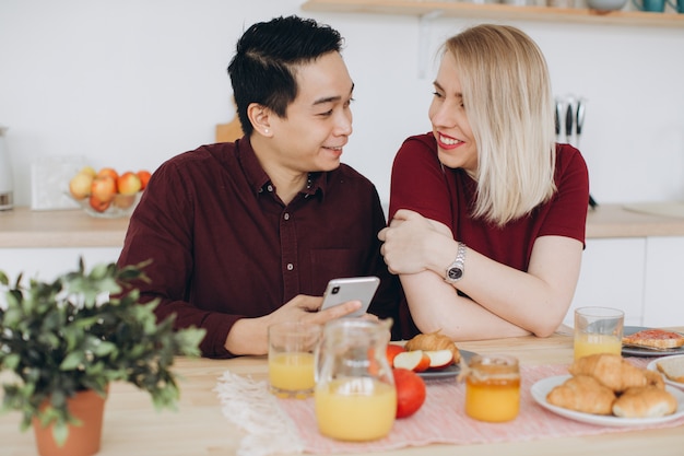 Asiatischer Mann und seine kaukasische blonde Frau frühstücken. Sie verbringen Zeit miteinander in der Küche und schauen sich Videos am Telefon an.