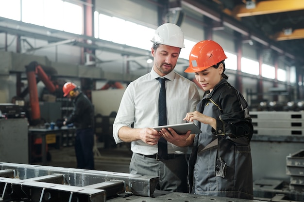 Asiatischer Metallbearbeitungsspezialist bespricht die technische Skizze mit dem Ingenieur, während er die technische App auf dem Tablet verwendet