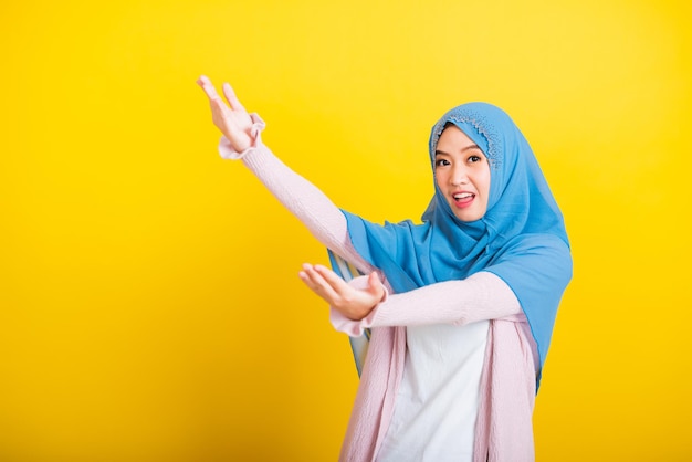 Asiatischer muslimischer Araber, Porträt einer glücklichen, schönen jungen Frau, Islam, religiöse Kleidung, Schleier, Hijab, lustiges Lächeln, sie öffnet die Arme für eine Umarmung oder hebt die Hand, um Gegenstände aufzuheben, erhält etwas von oben isoliertes Gelb