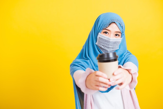 Asiatischer muslimischer Araber, Porträt einer glücklichen, schönen jungen Frau Islam, religiöse Kleidung, Schleier, Hijab und Gesichtsmaske, schützen sie unter Quarantäne, Coronavirus, Kaffee-Pappbecher, isolierter gelber Hintergrund