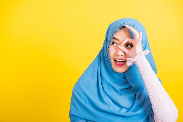 Asiatischer muslimischer Araber, Porträt einer glücklichen schönen jungen Frau Islam religiöser Kleidung Schleier Hijab lustiges Lächeln sie zeigt Gestenfinger in Ordnung Gestensymbol, OK-Schild über ihren Augen isoliert gelber Hintergrund