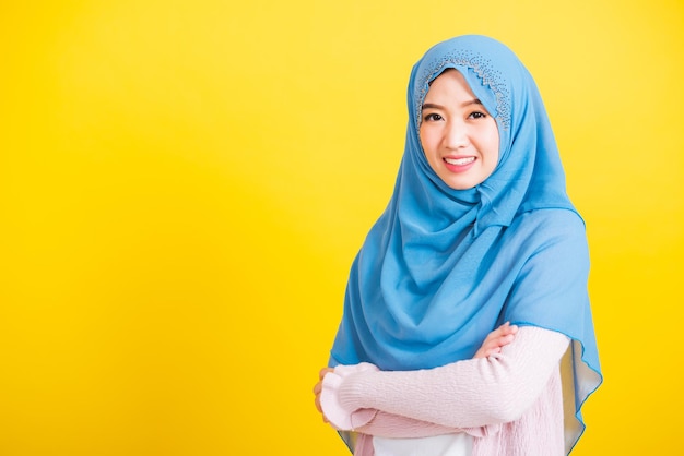 Asiatischer muslimischer Araber, Porträt einer glücklichen, schönen jungen Frau, islamische religiöse Kleidung, Schleier, Hijab, sie lächelt, steht mit gekreuzten Armen, Studioaufnahme isoliert auf gelbem Hintergrund