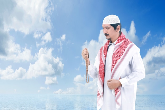 Asiatischer muslimischer Mann, der mit Gebetsperlen auf seinen Händen mit einem blauen Himmel betet