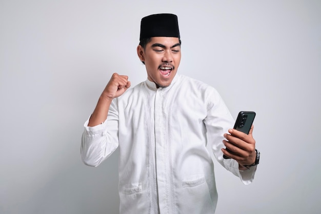 Foto asiatischer muslimischer mann in weißer kleidung mit smartphone und gewinnender geste mit handy