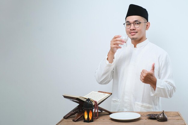 Asiatischer muslimischer Mann lächelt und Daumen hoch, während er ein Glas Wasser zum Fastenbrechen auf weißem Hintergrund trinkt