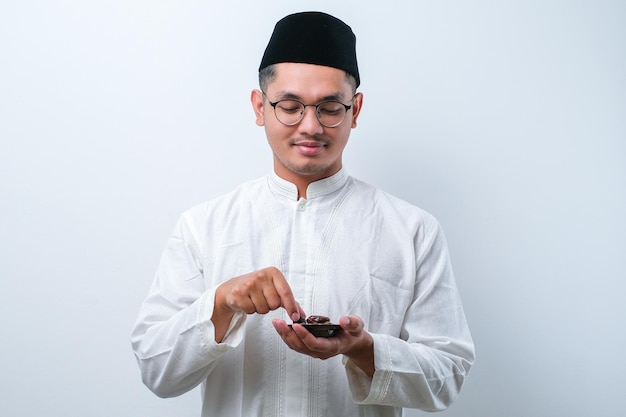 Asiatischer muslimischer Mann mit einer Schüssel mit Dattelfrüchten auf weißem Hintergrund, der zum Fastenbrechen des Iftar isst