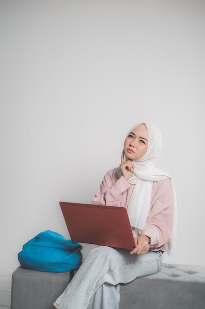 Asiatischer muslimischer Student, der Laptop vor weißem lokalisiertem Hintergrund hält, der oben sitzt und denkt