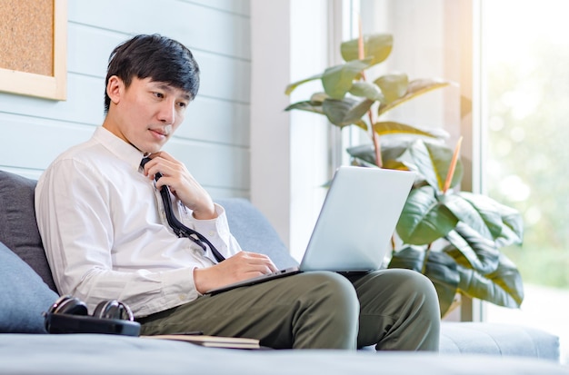 Asiatischer nachdenklicher junger gutaussehender professioneller erfolgreicher männlicher geschäftsmannangestellter im formellen geschäftsoutfit, der auf gemütlicher sofacouch sitzt und ideen denkt, die mit laptop-computer im wohnzimmer arbeiten.