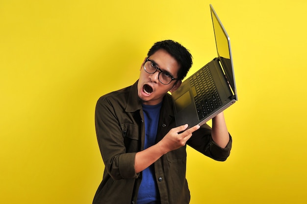 Asiatischer Online-Freiberufler sehr wütend vor seinem Laptop, bekommt einen schlechten Bericht vom Kunden, isoliert auf Gelb