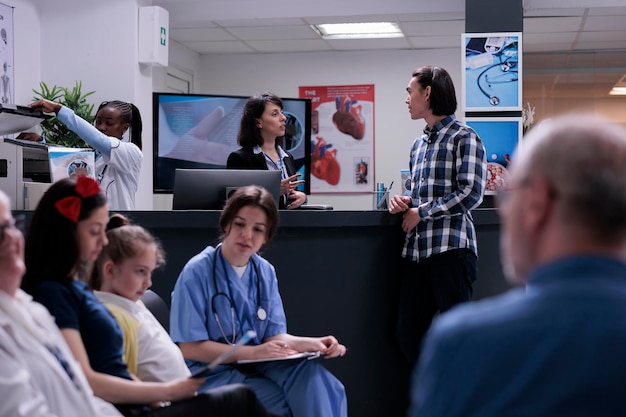 Asiatischer Patient spricht mit Krankenhausrezeptionistin, die an der Rezeption steht, während Krankenschwester junges Mädchen im Wartezimmer untersucht. Junger Mann, der mit verschiedenen Patienten an einem Arzttermin in einer Klinik teilnimmt.