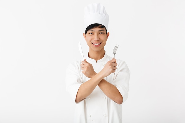 asiatischer positiver Hauptmann in der weißen Kochuniform, die an der Kamera lächelt, während Besteck lokalisiert über weißer Wand hält