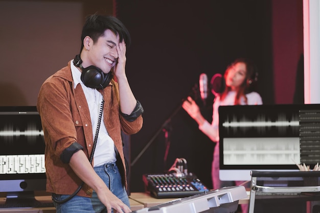 Asiatischer Produzent, der Tonmischpult bereitsteht Glücklicher männlicher Musikkomponist-Künstler mit einer Sängerin im Hintergrund