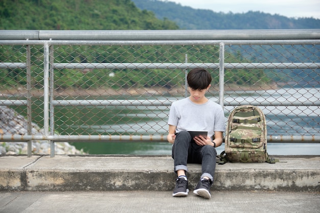 Asiatischer Reisender, der Tablette auf der Brücke über dem Fluss, mit einem Rucksack gesetzt auf die Seite sitzt und verwendet. - Bild