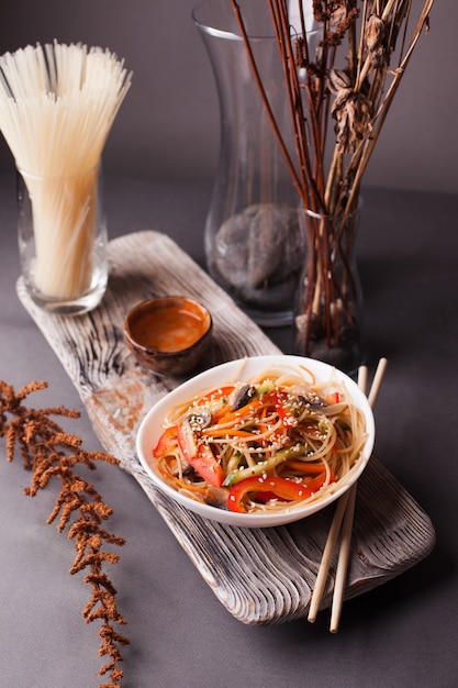 Asiatischer Salat mit den Nudeln und Gemüse, besprüht mit Samen des indischen Sesams mit Essstäbchen auf einem hölzernen Behälter. Asiatische Küche, Beilage, gesundes Essen, vegetarisches Menü.