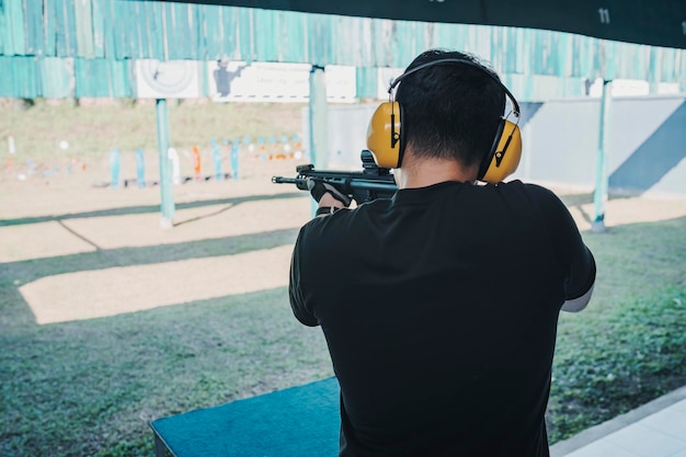 Asiatischer Schütze mit geräuschunterdrückenden Kopfhörern und schwarzer Kleidung, der auf dem Schießstand Schießgewehr übt Schießsport für Meditation und Selbstverteidigung Freizeitaktivitäten