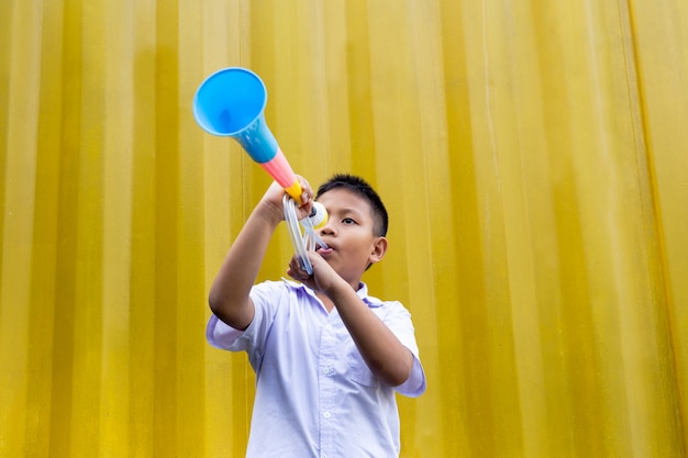 Asiatischer Schuljunge, der ein buntes Horn im gelben Hintergrund durchbrennt.