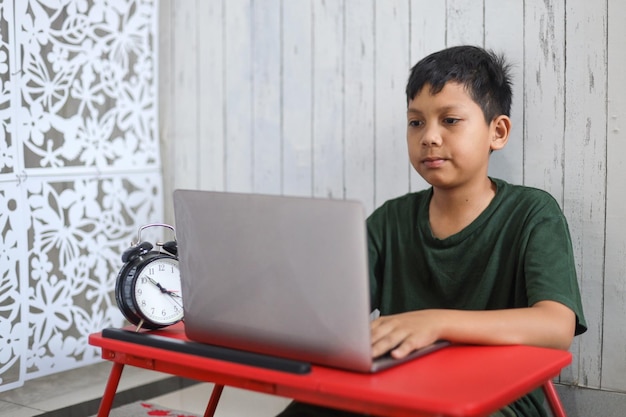 Asiatischer Schuljunge, der zu Hause mit einem Laptop lernt und online mit dem Videoanruflehrer zu Hause lernt.