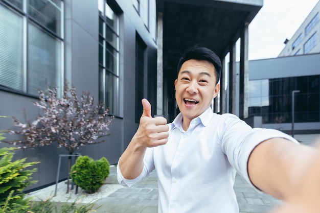 Asiatischer Student macht Selfie, schaut auf Telefonbildschirm und lächelt Videoanruf mit Freunden