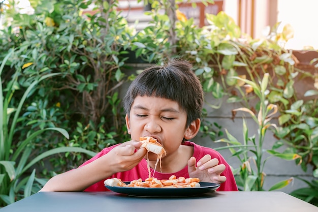 Asiatischer süßer Junge im roten Hemd, der köstlich und glücklich Pizza isst.