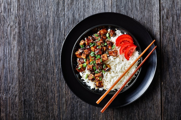 Asiatischer Teriyaki-Tofu mit Reis, bestreut mit Frühlingszwiebeln, Sesam, serviert mit Reis, Tomatenscheiben, auf einer schwarzen Schüssel mit Stäbchen auf dunkler Holzoberfläche, Draufsicht, flache Lage, Nahaufnahme