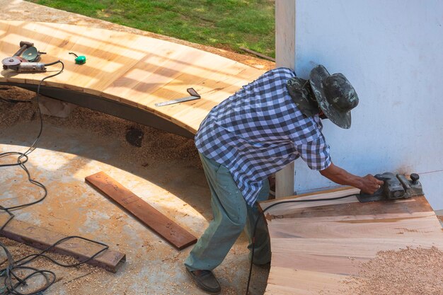 Asiatischer Tischler, der elektrische Hobel verwendet, um gekrümmte Holzbankoberflächen in der Werkstatt im Freien zu rasieren