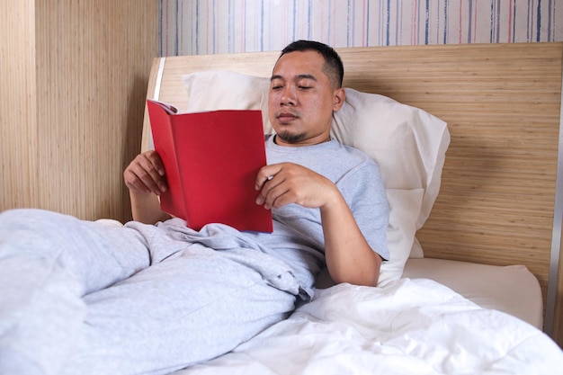 Asiatischer Typ liest ein Buch, das zu Hause im bequemen Bett im Schlafzimmer liegt. Mann genießt Longread-Fiktion