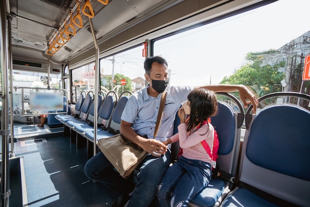 Asiatischer Vater bringt seine Tochter zur Schule, indem er mit öffentlichen Verkehrsmitteln mit dem Bus fährt und eine Gesichtsmaske trägt