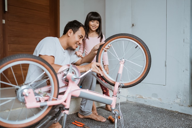 Asiatischer Vater, der Räder hält, während er mit seiner Tochter Fahrrad zusammenbaut