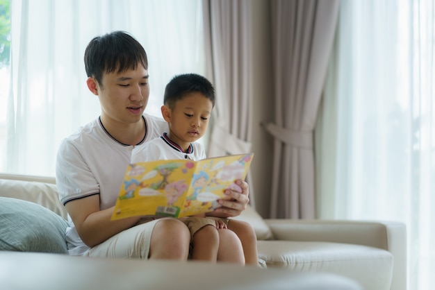 Asiatischer Vater hält ein Buch, um seinen Kindern das Lesen auf dem Sofa im Wohnzimmer beizubringen, Väter interagieren den ganzen Tag mit ihren Kindern.