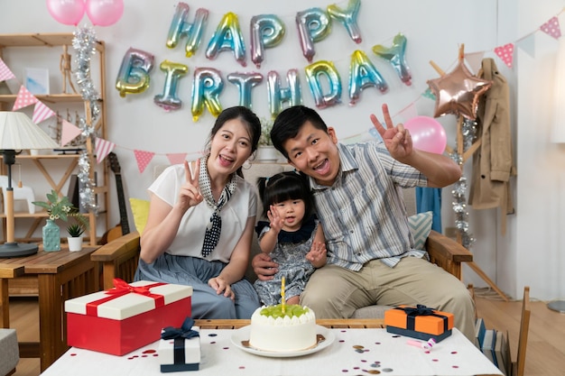 asiatischer vater, mutter und kleines mädchen lächeln mit friedenshandzeichen in die kamera, während sie geburtstag in einem gemütlichen wohninneren mit bunter partydekoration feiern