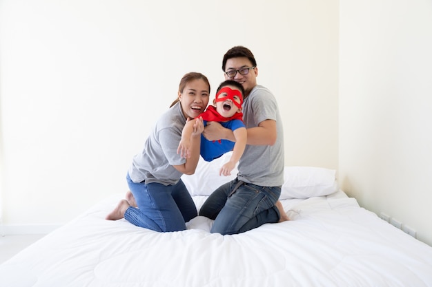 Asiatischer Vater, Mutter und Sohn spielen Superhelden auf dem Bett im Schlafzimmer. Freundliche Familie, die Spaß hat