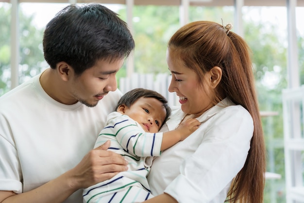 Asiatischer Vater und Mutter umarmen 9 Monate kleinen Sohn auf Sofa. Sie lächeln und berühren das Baby mit Liebe im Wohnzimmer zu Hause