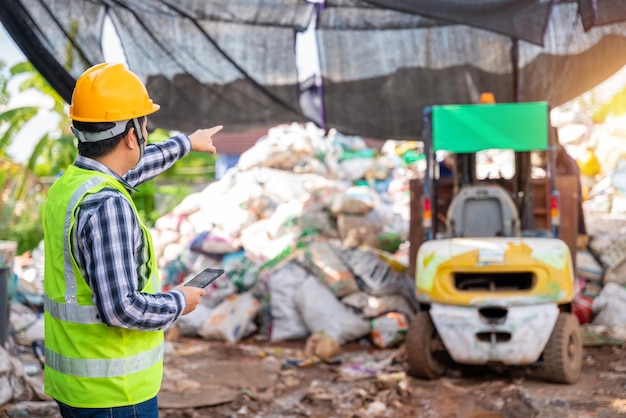 Asiatischer vorarbeiter, der in einer recyclingfabrik arbeitet und ein tablet in der hand hält und die abfallfabrik mit einem gabelstapler im hintergrund betrachtet