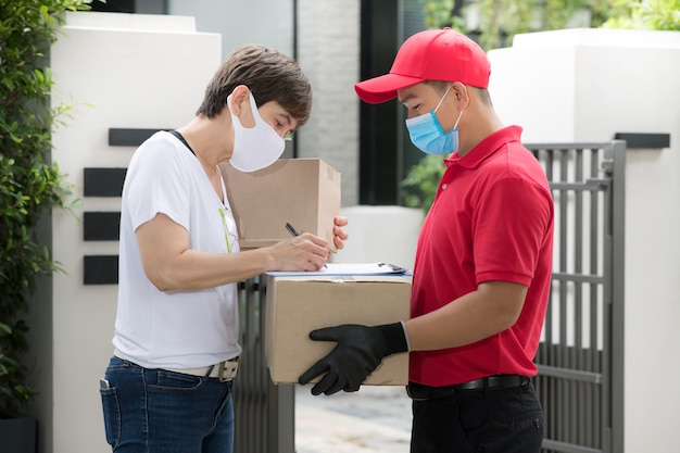 Asiatischer zusteller, der gesichtsmaske und handschuhe in der roten uniform trägt, die paketbox zum empfänger liefert