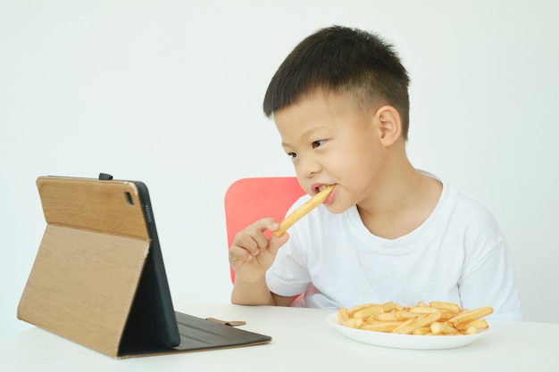 Asiatisches 5-jähriges kleines Kindergartenjungenkind, das Pommes frites isst, während es sich ein Video vom Tablet-PC ansieht, das über weißem Wandhintergrund isoliert ist Ungesunde Kindernahrung Gadgetsüchtiges Kinderkonzept