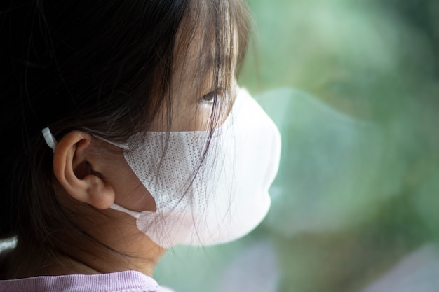 Asiatisches 6 oder 7 Jahre altes Kind mit medizinischer Maske. Kleines Mädchen, das am Fenster steht und nach draußen schaut. Sie sieht traurig und gelangweilt aus. Sie kann krank sein oder unter Quarantäne der Coronavirus-Krankheit 19 (COVID-19).