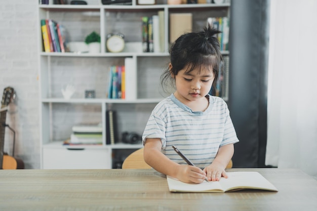 Asiatisches Baby, das ein blau gestreiftes Hemd trägt, schreibt Notizen in ein Notizbuch, um online auf einem Holztisch im Wohnzimmer zu Hause zu lernen. Bildung lernt online von zu Hause aus