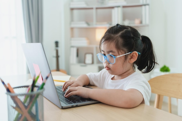 Asiatisches Baby, das ein weißes T-Shirt und eine Brille trägt, benutzt einen Laptop und lernt online auf einem Holztisch im Wohnzimmer zu Hause. Bildung lernt online von zu Hause aus