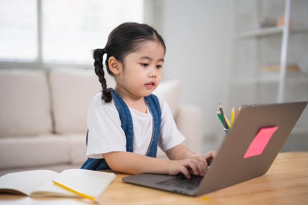 Asiatisches Babymädchen lächelt, benutzt Laptop oder schreibt, zeichnet Farbe auf Notizbuch, lernt online auf Holztisch, Schreibtisch im Wohnzimmer zu Hause. Bildung lernt online von zu Hause aus